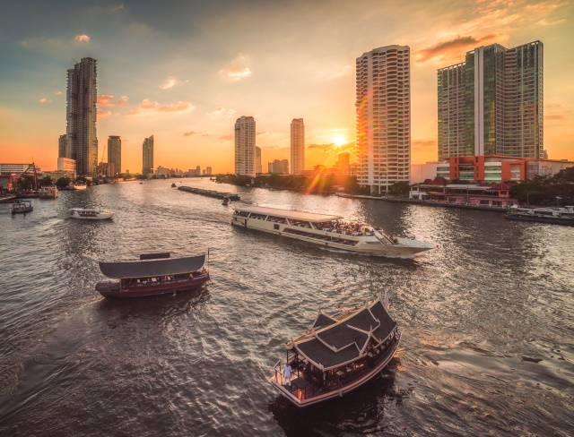 Busy Chao Phraya River With Passenger Boats And Skyscrapers At Sunset As Seen From Taksin Bridge In Bangkok, Thailand