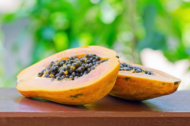 A Papaya Fruit Cut In Half Laying On Wood With Trees In The Background